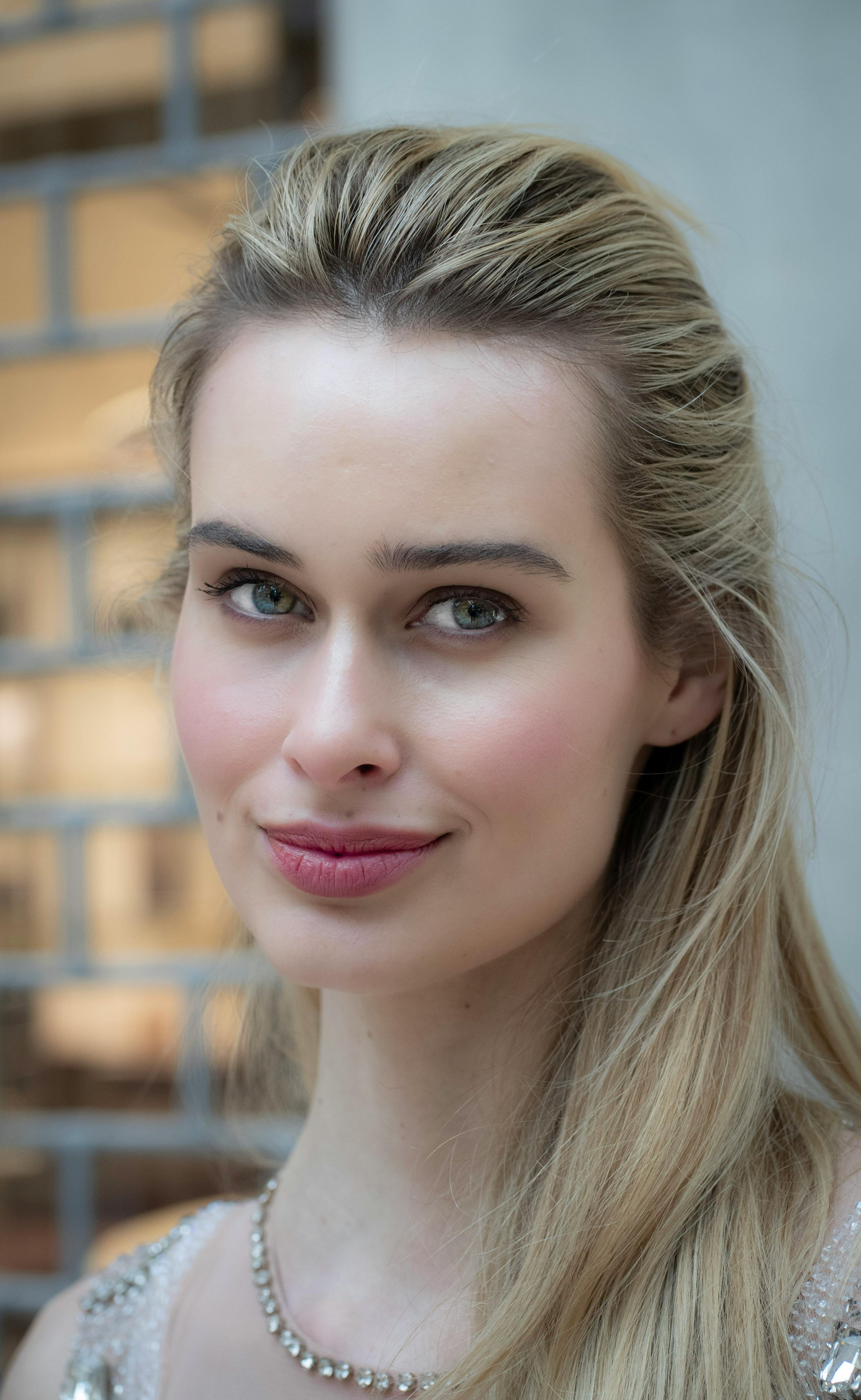 Close-up portrait of a woman with long blonde hair and striking green eyes, wearing a subtle smile, adorned with a delicate necklace, set against a softly blurred background.