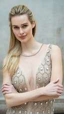 A woman with long blonde hair poses confidently, wearing a sparkling, intricately beaded dress with a deep neckline, against a neutral gray background.