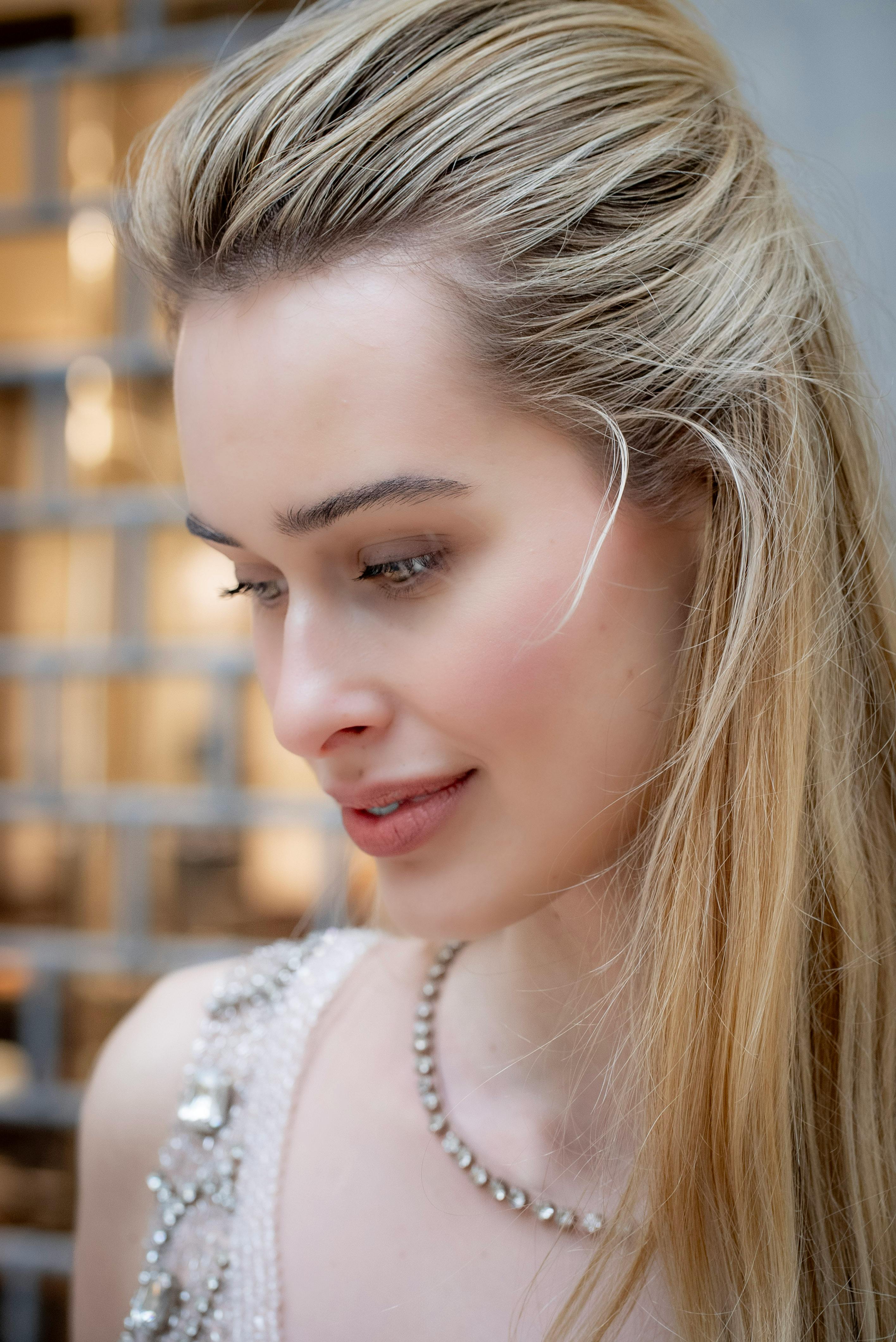Close-up of a young woman with long blonde hair, wearing a beaded dress and a sparkling necklace, smiling softly while looking downward.