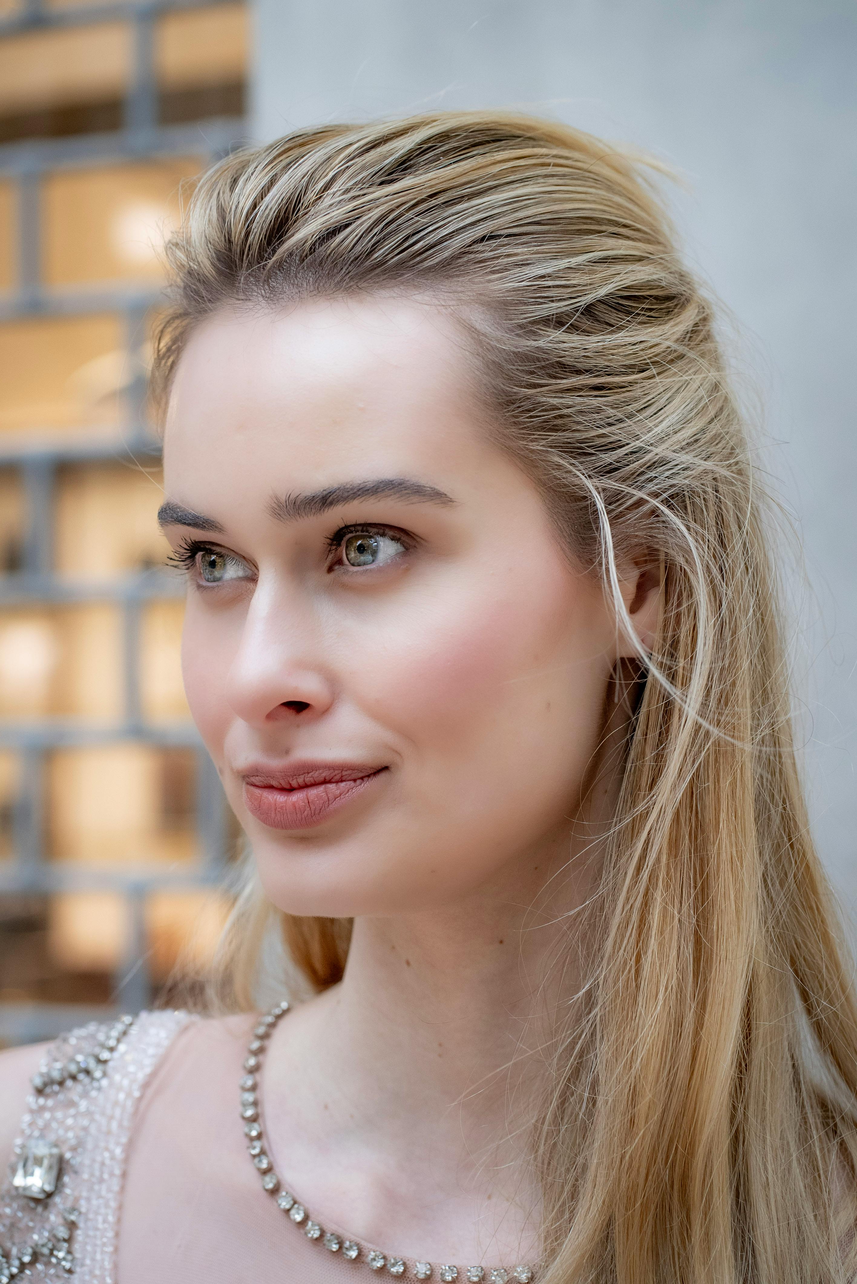 Portrait of a young woman with long blonde hair and green eyes, wearing a beaded dress, with a soft smile against a blurred background.