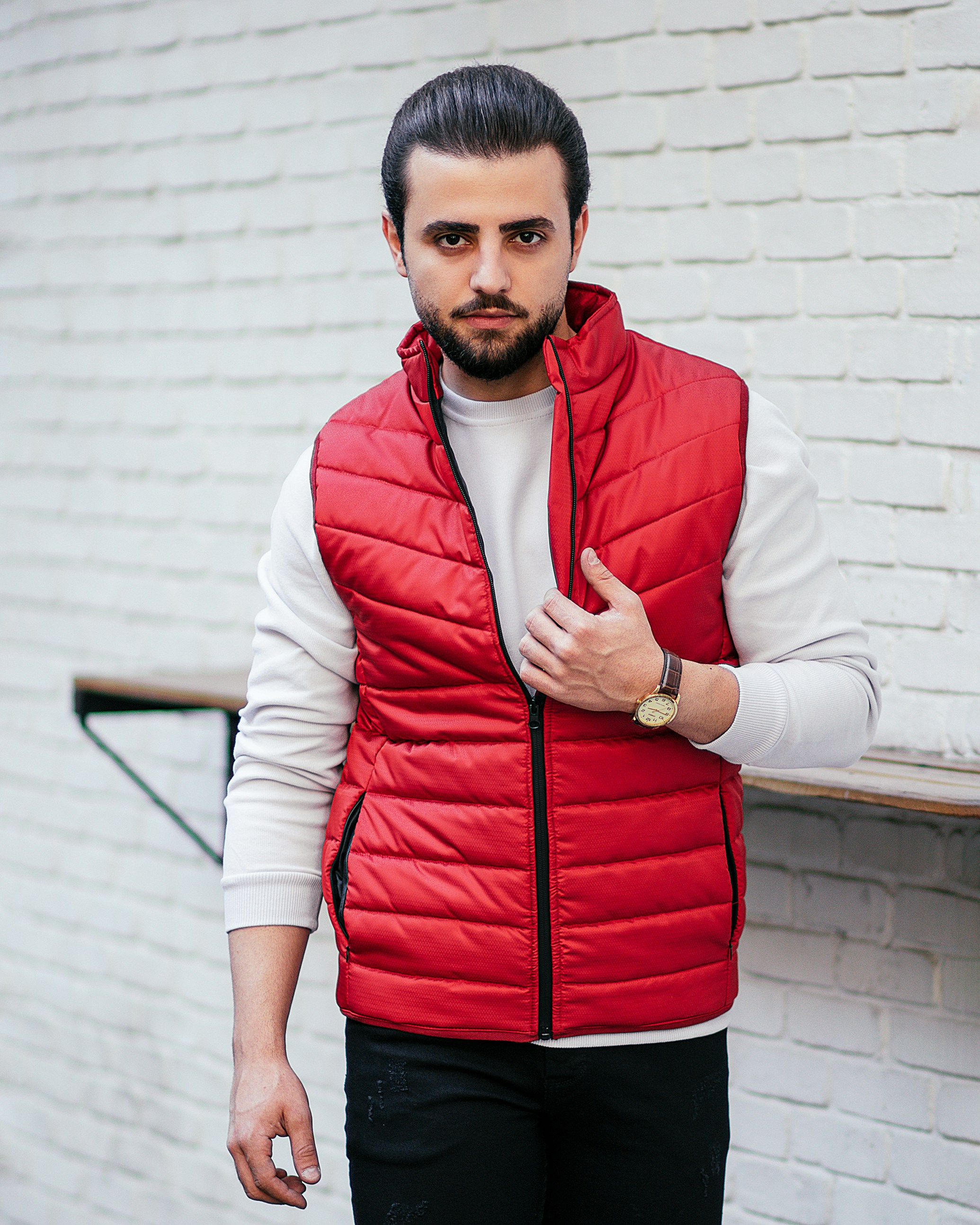 Man wearing a red puffer vest over a white sweater, standing against a white brick wall, showcasing a stylish casual look.