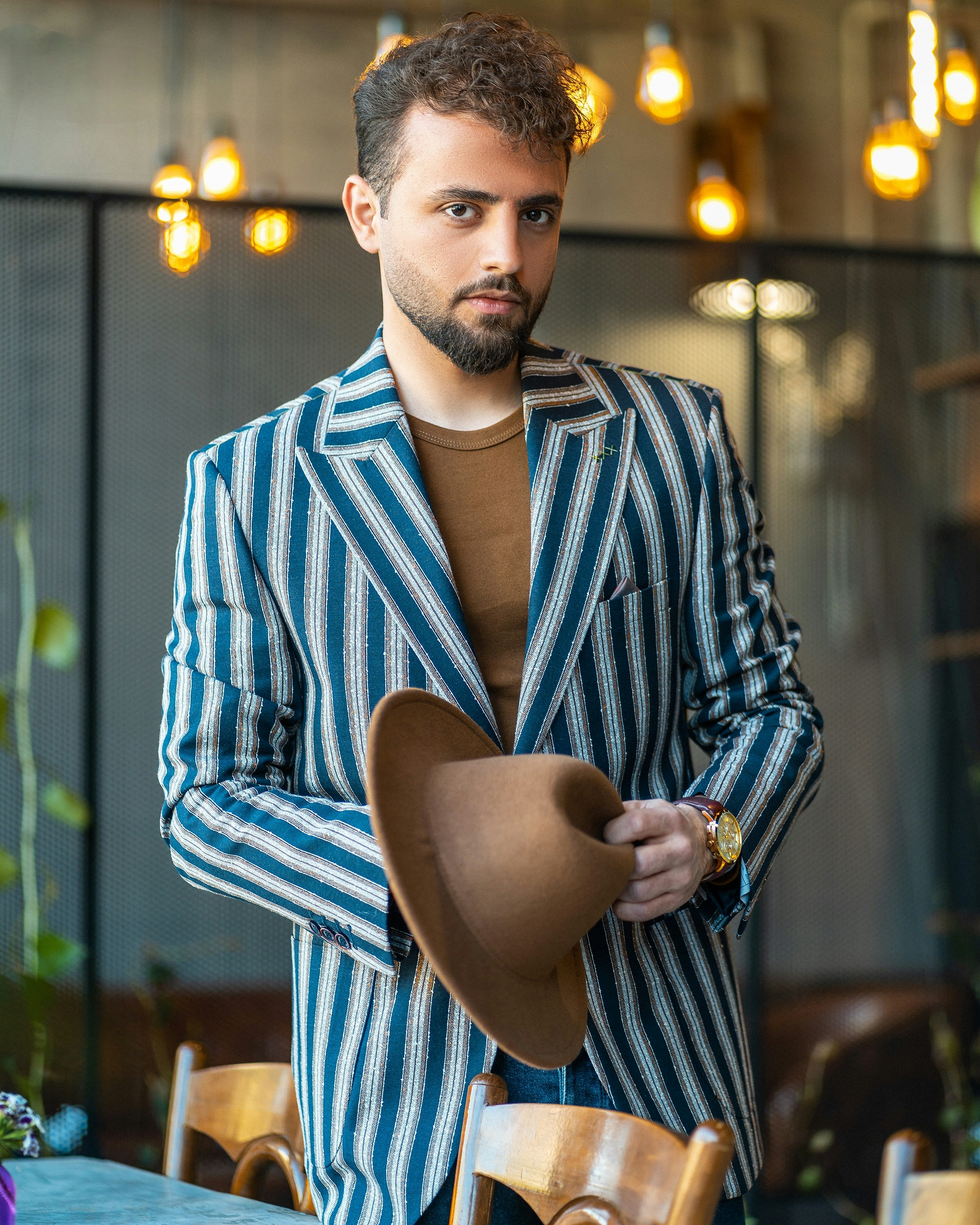 Stylish man in a striped blazer holding a brown hat, set in a modern café with warm lighting.