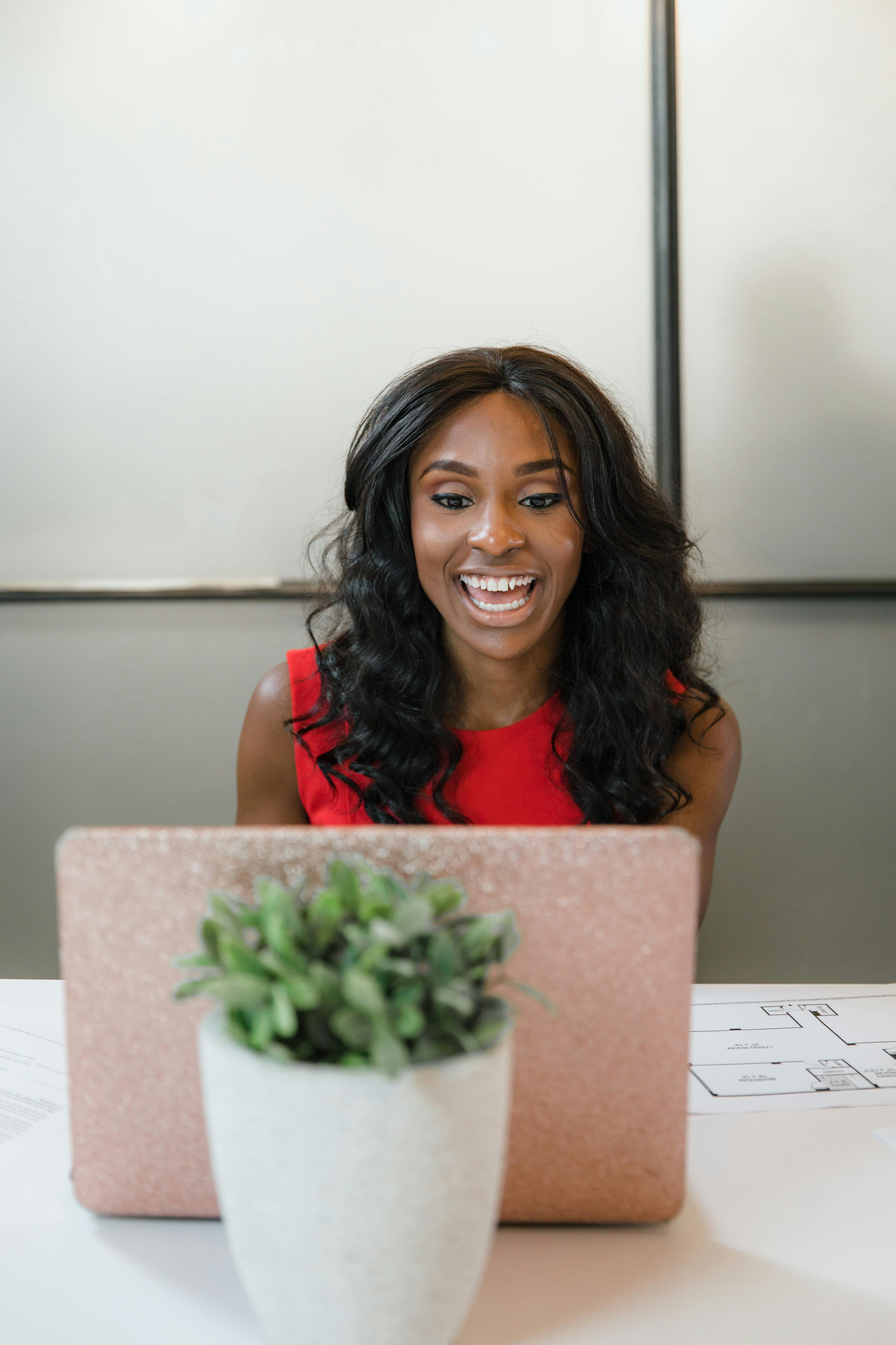 Smiling young woman in a red dress sitting at a desk with a laptop and a potted plant, engaged in work or a video call.