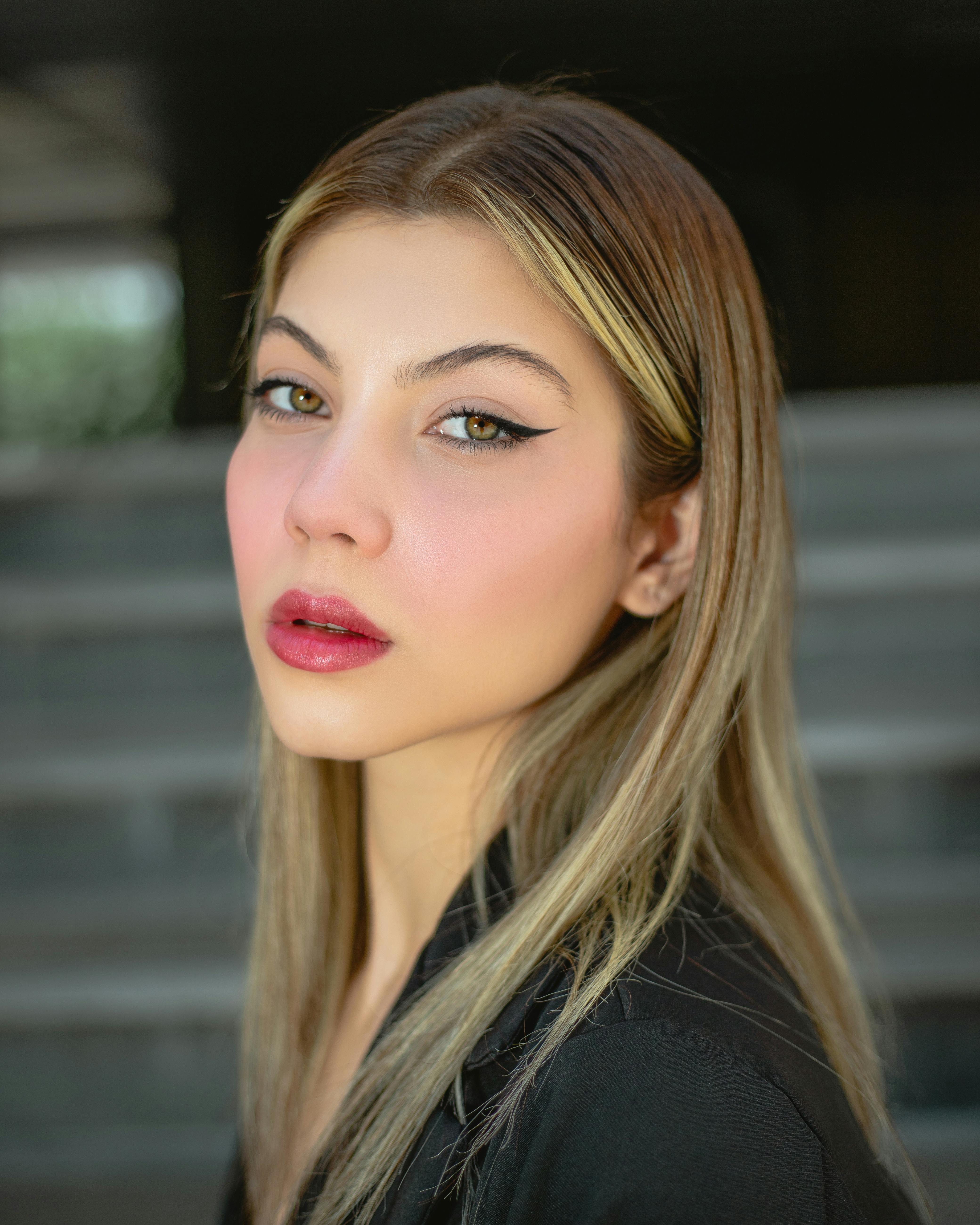 Portrait of a young woman with long, straight blonde hair and striking green eyes, featuring bold winged eyeliner and soft pink lips, set against a blurred outdoor background.