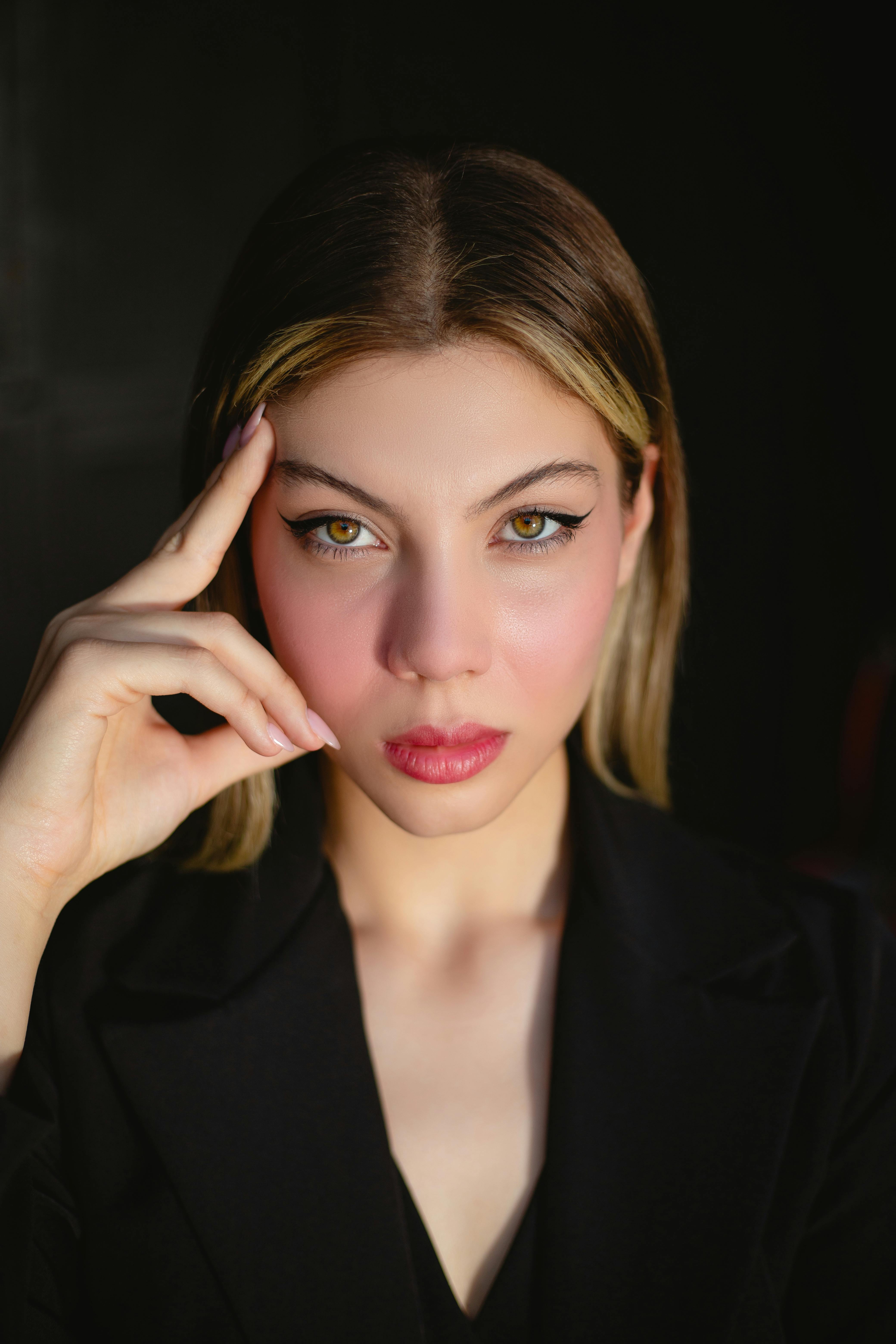 Portrait of a young woman with blonde highlights, showcasing bold eyeliner and soft pink lip color, thoughtfully resting her chin on her hand against a dark background.