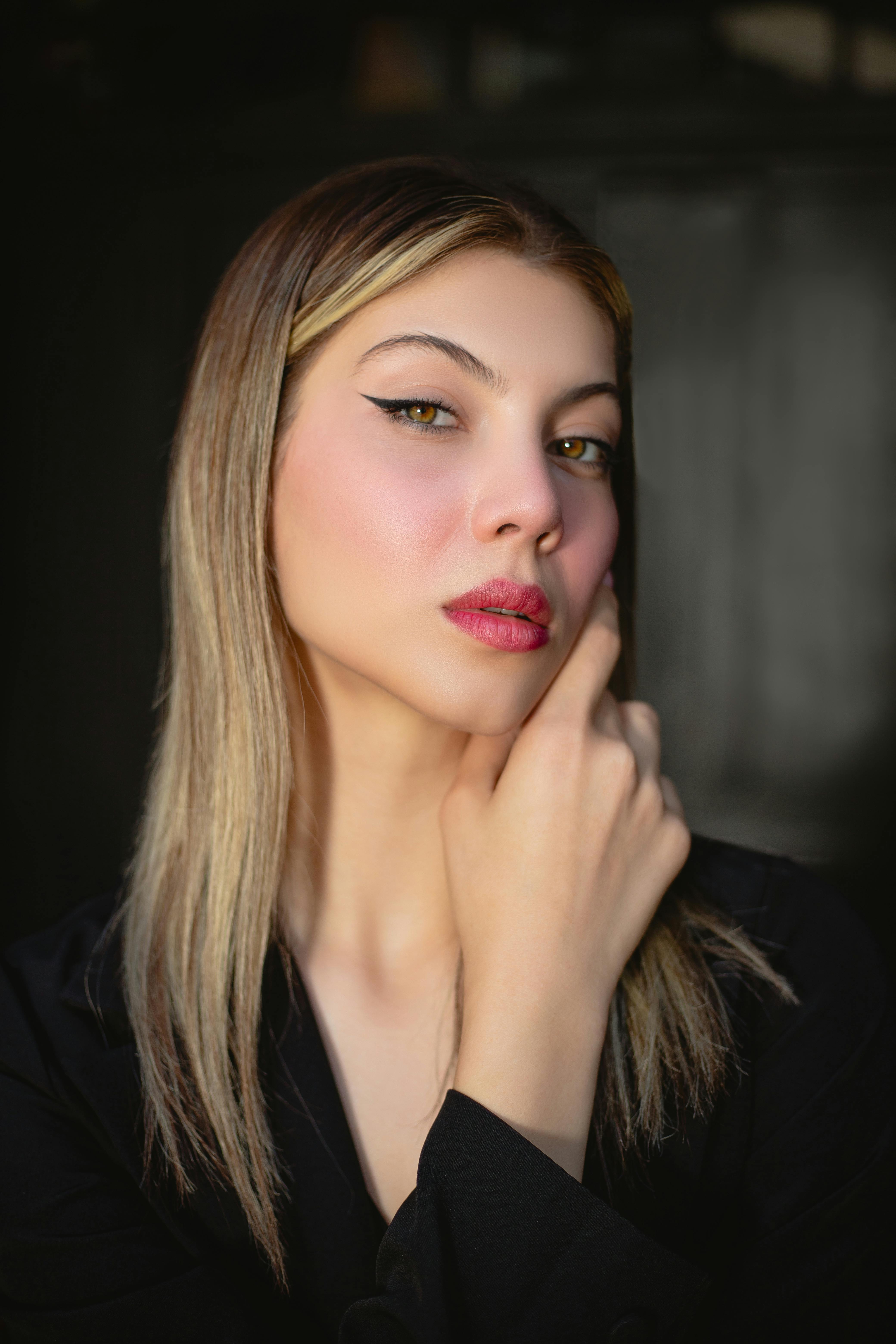 Portrait of a young woman with long, straight hair and striking green eyes, wearing bold makeup and a black outfit, against a dark backdrop.