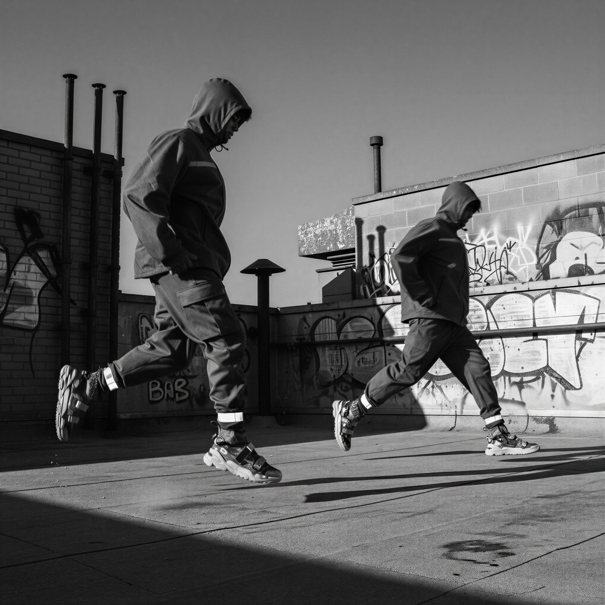 Streetwear editorial style photography, gritty urban rooftop setting, harsh directional sunlight creating deep shadows, high-contrast monochrome palette, oversized silhouettes, technical fabrics, graffiti backdrop, dynamic motion blur, sneaker culture influence, urban edge mood, high fashion magazine quality composition
