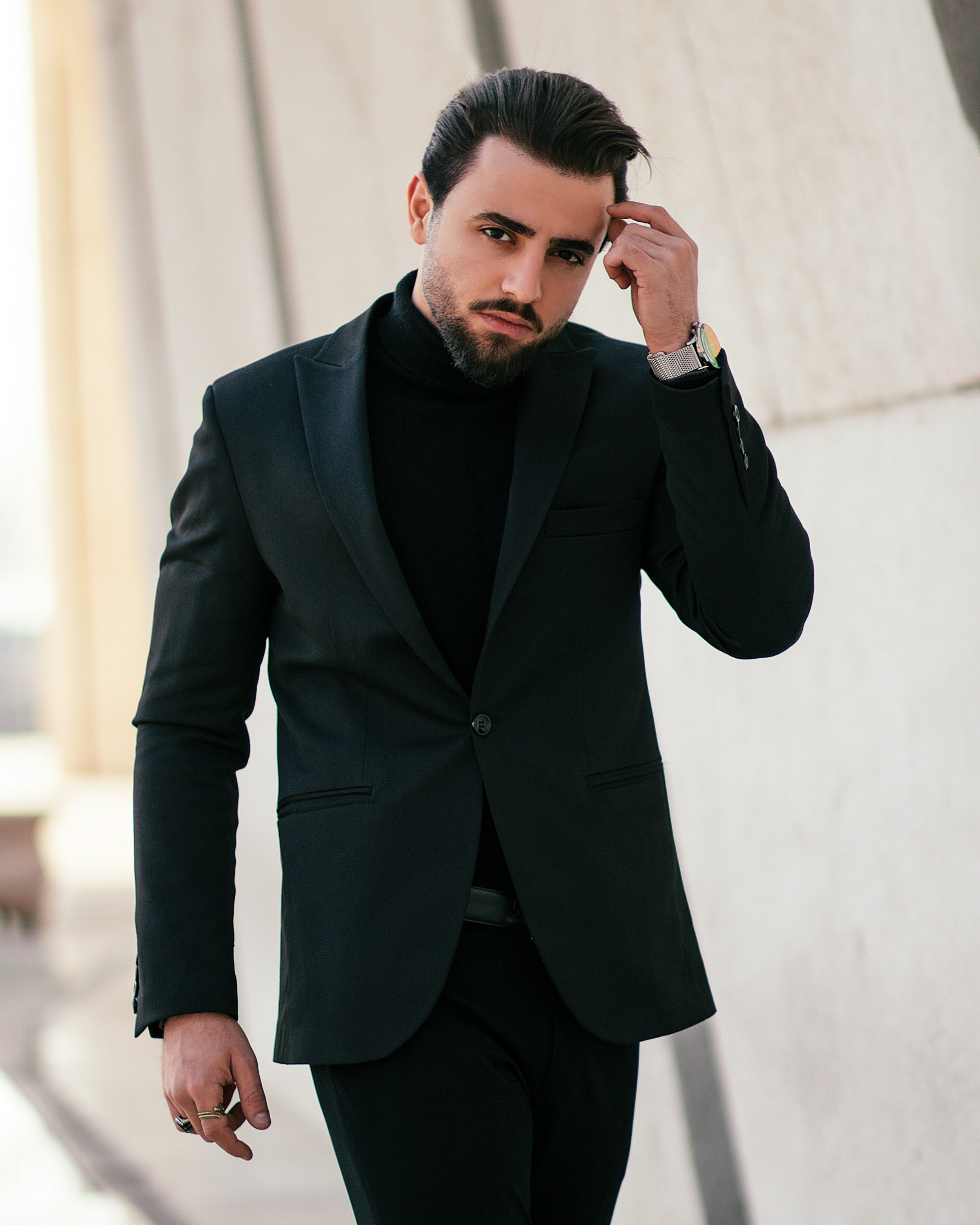 Stylish man in a black suit and turtleneck walks confidently, adjusting his hair against a modern architectural backdrop.