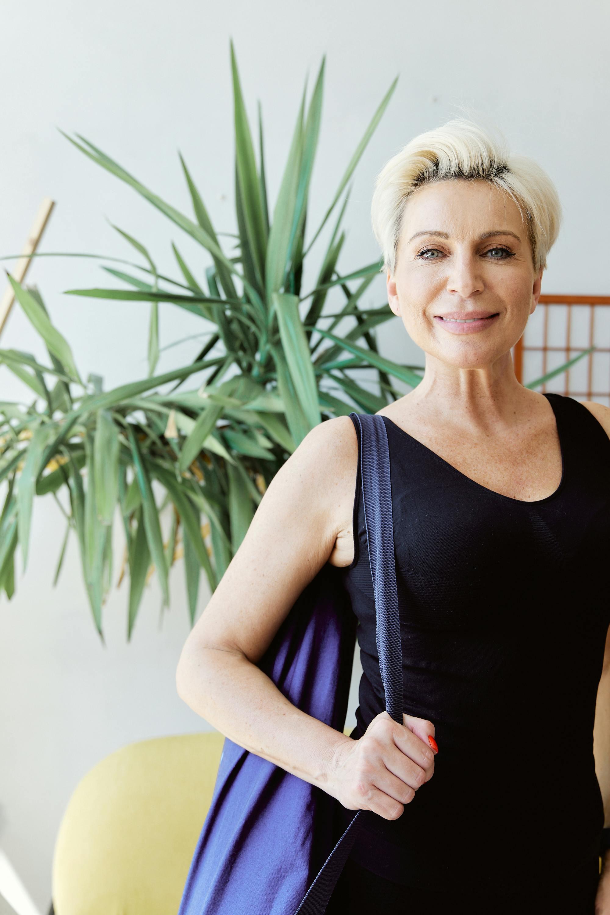 Smiling middle-aged woman with short blonde hair in black top carrying a purple yoga mat bag indoors.