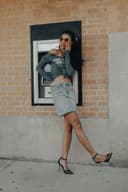 Fashionable young woman posing confidently in front of an ATM, wearing a grey off-the-shoulder top, a denim skirt, and black high heels, with sunglasses accentuating her stylish look.