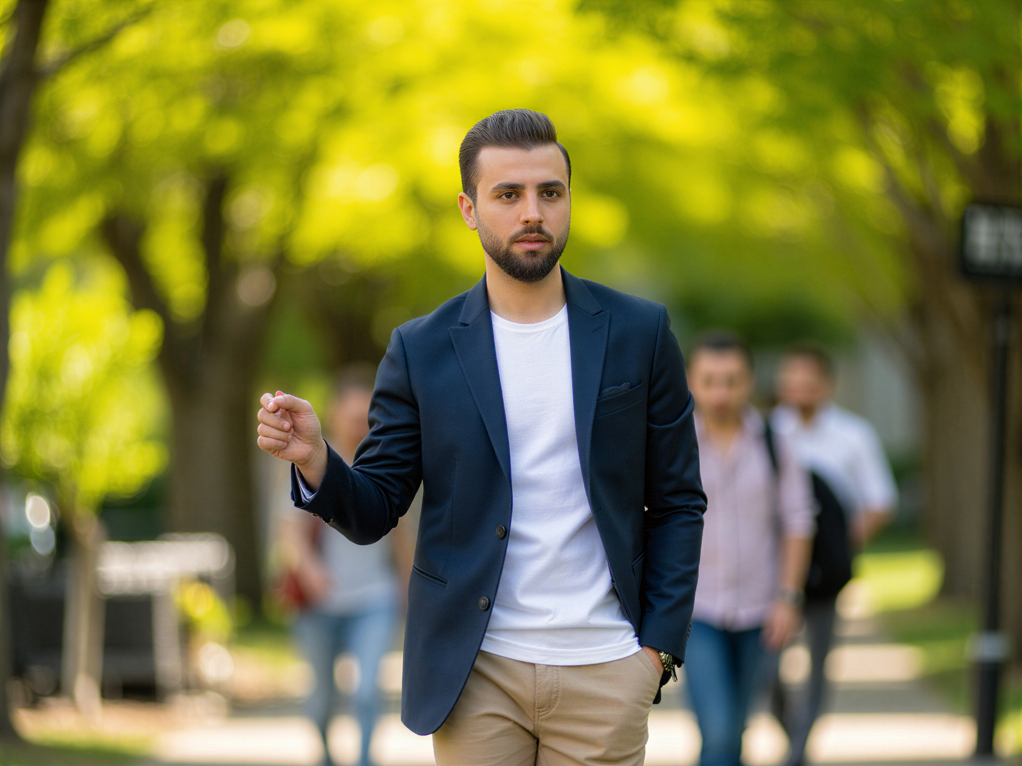 A charismatic male, OD62S, aged 32, captured on a vibrant college campus in full bloom. He stands casually against a sunlit backdrop of lush greenery, wearing a tailored navy blazer over a crisp white tee and relaxed chinos. His joyful expression radiates warmth and approachability, making him the embodiment of youthful exuberance. The pose is dynamic, with one hand tucked casually in his pocket while the other gestures animatedly, as if sharing a story with friends. The composition highlights the lively atmosphere of campus life, with fellow students blurred in the background, exuding a sense of camaraderie and happiness that envelops the scene.