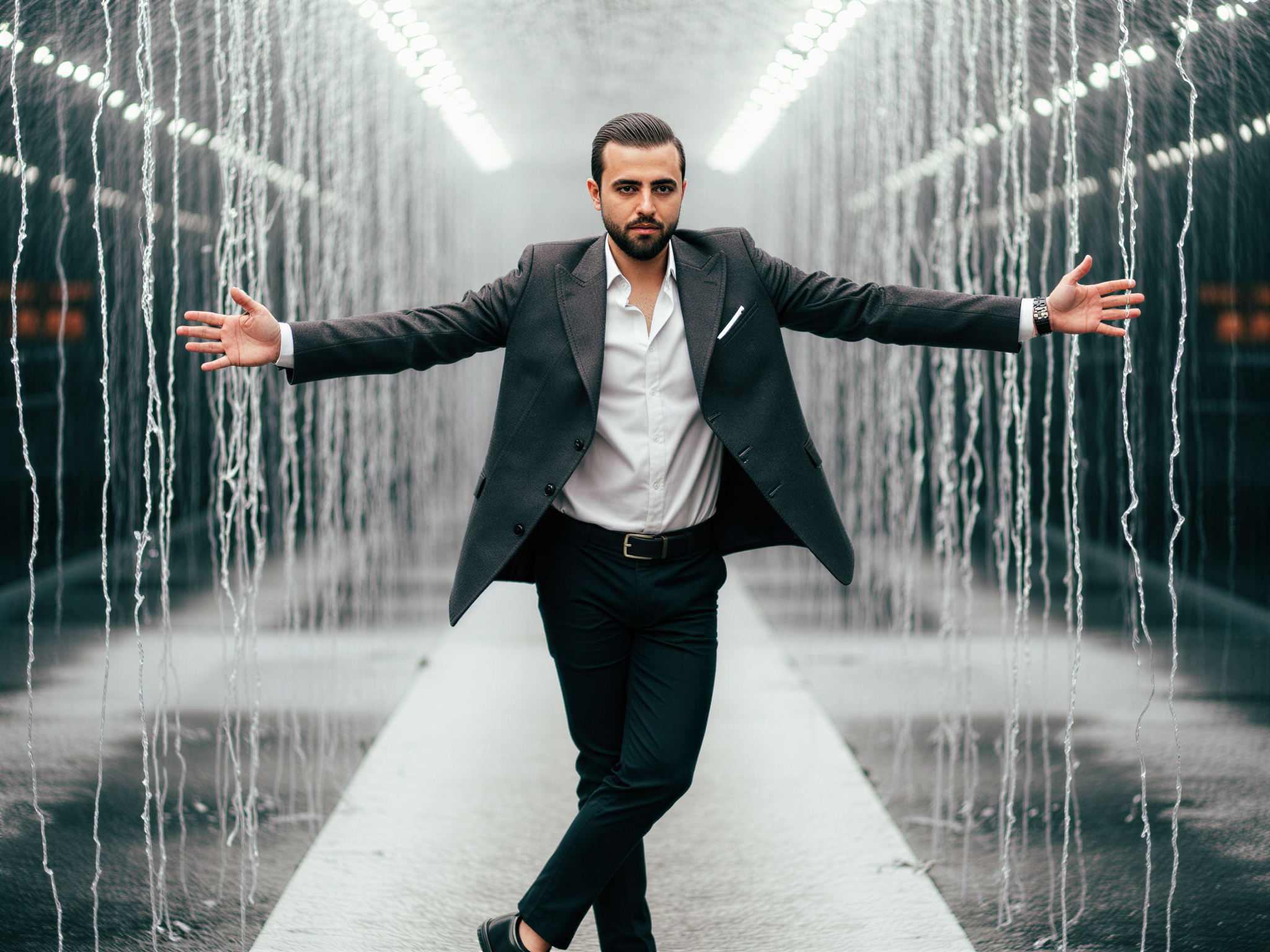 A 32-year-old male, OD62S, stands joyfully in a Rain Room installation, where the environment combines art and technology with cascading water dripping around him. He wears a tailored, charcoal trench coat over a crisp white shirt, complemented by smart trousers and polished black shoes. His arms are outstretched, and his face beams with happiness as the water forms a curtain, creating a mesmerizing interplay of light and droplets. The atmosphere is playful and surreal, evoking a sense of freedom and joy in this innovative space. The lighting subtly highlights his features, while the reflective surfaces of the room create an enchanting, immersive experience.