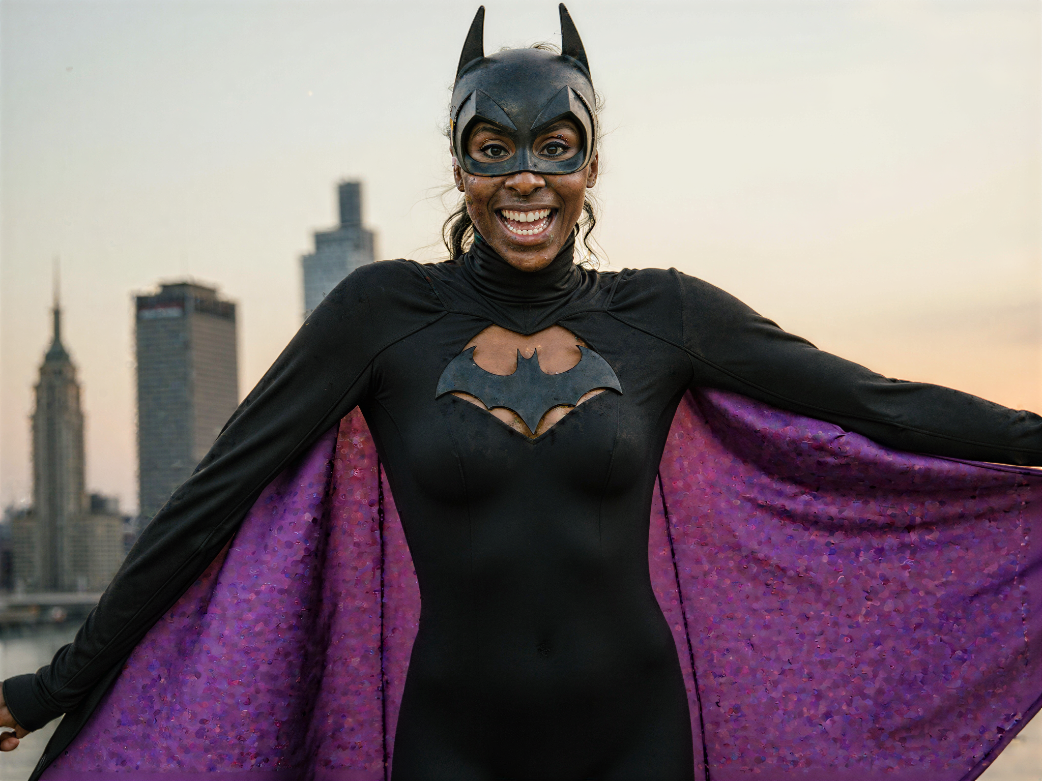 A joyful female figure, OD62S, aged 28, embracing the spirit of a superhero in a realistic interpretation of Batman. She wears a tailored black bodysuit made from matte neoprene, adorned with subtle textures and curved seams that enhance her athletic silhouette. A cape flows dramatically behind her, its inner lining a vibrant shade of purple that catches the light playfully. Her hair is styled into a sleek high ponytail, and she dons a playful version of the iconic Batman mask, revealing a bright, radiant smile that juxtaposes the character's usual seriousness. The setting captures an urban backdrop with skyline silhouettes, complemented by soft, warm lighting that evokes a sense of happiness and adventure. The mood is light and energetic, a celebration of strength and femininity while embodying the essence of a beloved comic book hero.