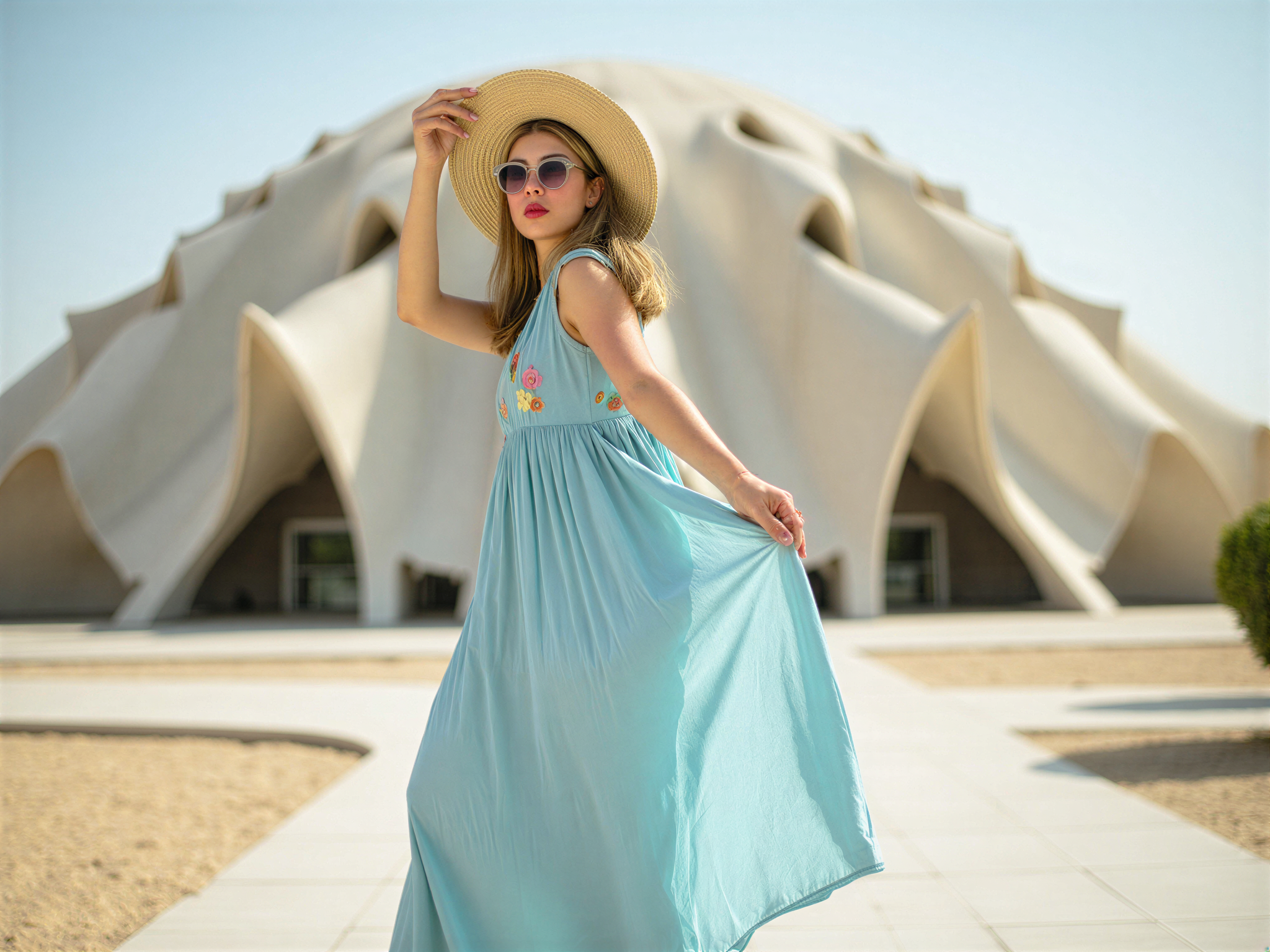 A joyful female figure, OD62S, aged 28, is standing amidst the stunning architecture of the Louvre Abu Dhabi. She wears a flowing, sky-blue silk maxi dress that moves gracefully with her, capturing the essence of flow and light. The dress features delicate, hand-embroidered floral details, symbolizing joy and life. Her hair is styled in loose waves that dance in the gentle breeze, complemented by oversized sunglasses and a wide-brimmed straw hat, giving her a carefree, chic summer vibe. The backdrop showcases the museum's iconic dome structure, casting intricate shadows, while natural sunlight bathes the scene in a warm glow, enhancing the mood of happiness and serenity. The image is framed to highlight both the subject and the architectural marvel, creating a harmonious blend of art and fashion.
